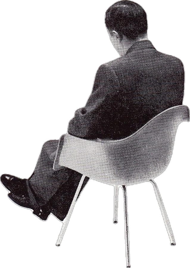 A man in a dark suit seated in a molded shell armchair, seen from behind.